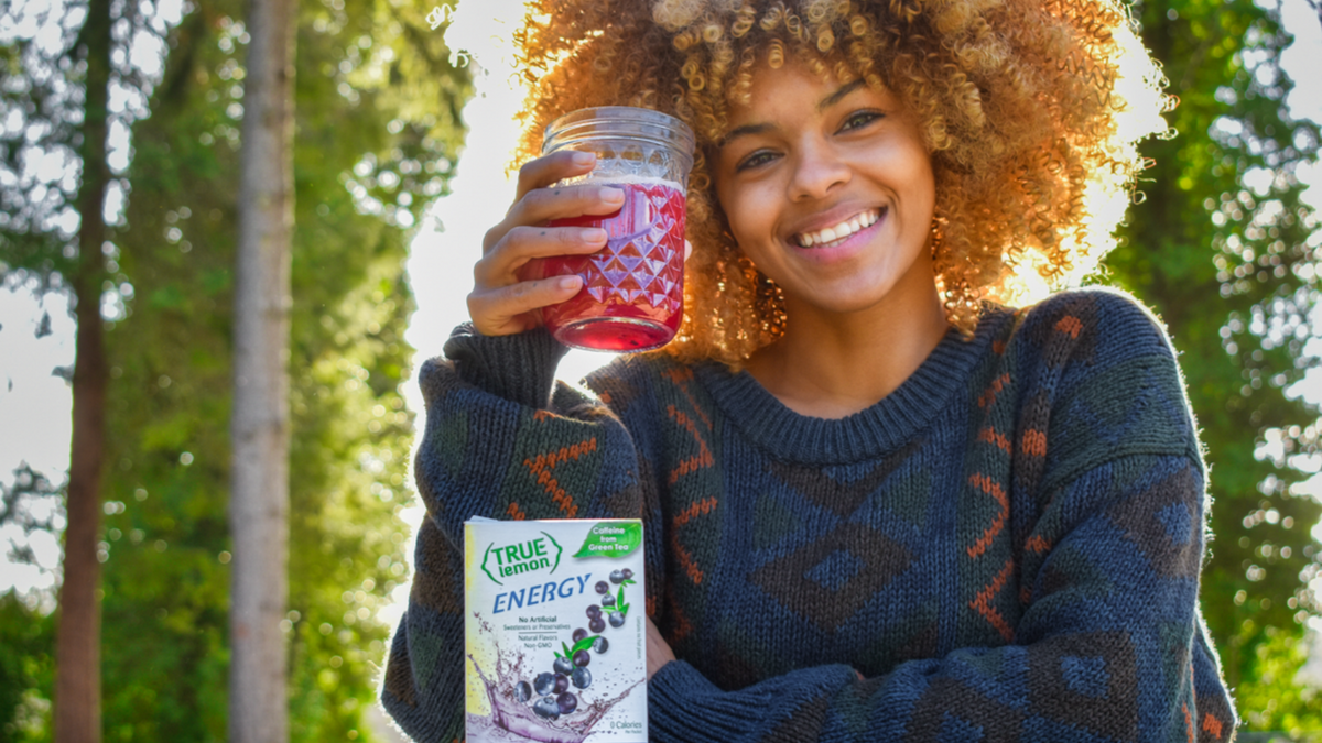 A young woman sits outside in the sun and drinks True Lemon Energy Blueberry Acai, made with Sustained Caffeine from green tea.