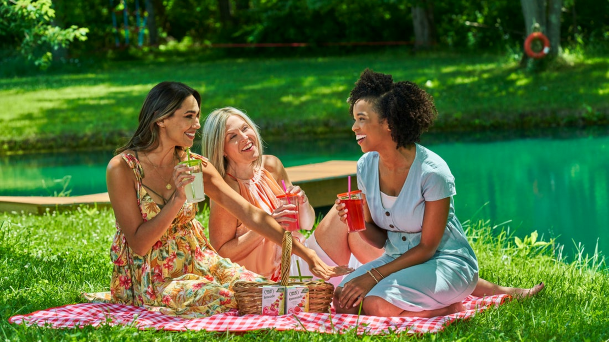 Three women sit on a picnic blanket in a park. They are laughing and drinking True Lemon Original Lemonade.